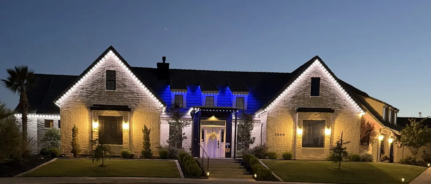 White brick ranch home lit with warm white roofline lighting and a deep blue accent zone over the central entry