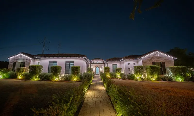 Spanish-style single-story home at night with crisp white permanent roofline lighting and green landscape uplighting on hedges